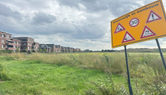 Op de voorgrond een bord met de waarschuwing 'wijk in aanbouw', op de achtergrond een leeg weiland met ernaast een woonwijk