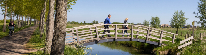 Man en vrouw wandelen over een houten brug van fietspad naar weiland