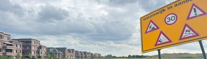 Op de voorgrond een bord met de waarschuwing 'wijk in aanbouw', op de achtergrond een leeg weiland met ernaast een woonwijk