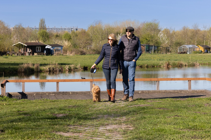 Man en vrouw met hond wandelen langs het water in Noorderpark-Ruigenhoek, met op de achtergrond flats in Utrecht Overvecht