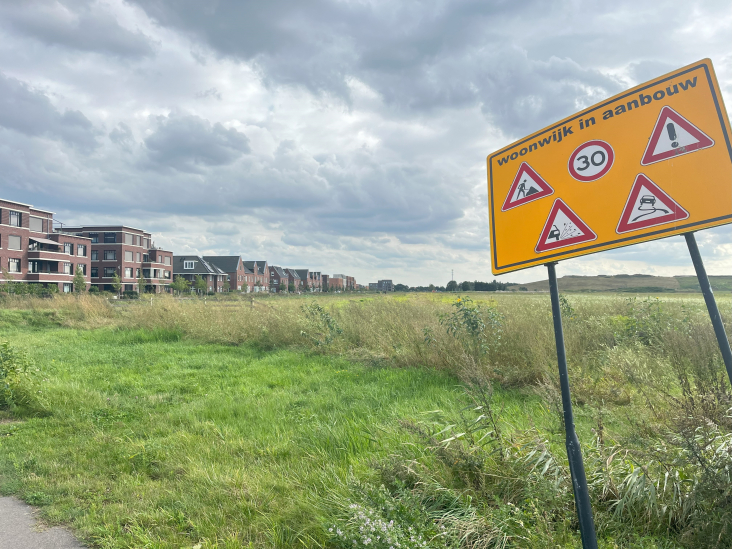 Op de voorgrond een bord met de waarschuwing 'wijk in aanbouw', op de achtergrond een leeg weiland met ernaast een woonwijk