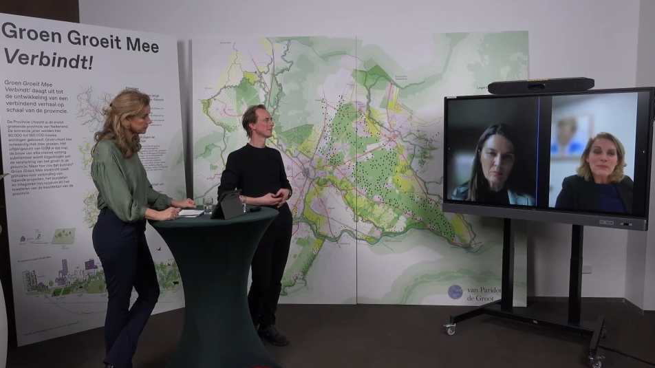 Foto van de studiosetting met de gespreksleider en spreker, met op een groot scherm twee gespreksdeelnemers die online aansluiten