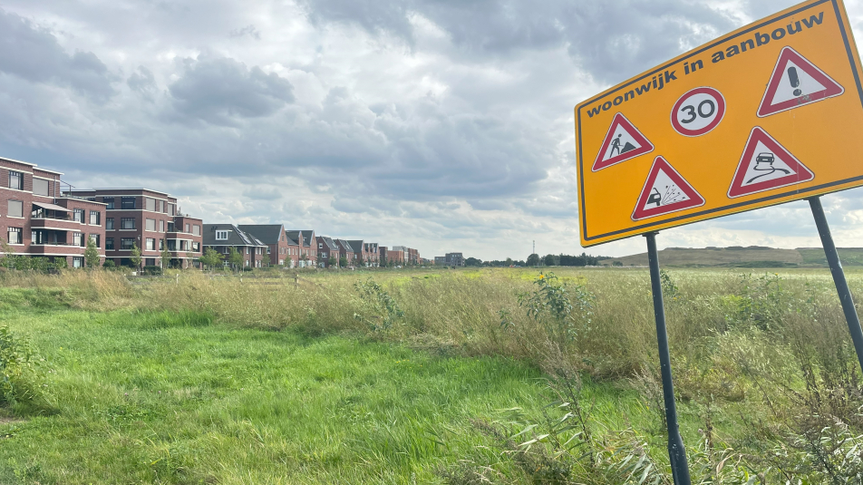 Op de voorgrond een bord met de waarschuwing 'wijk in aanbouw', op de achtergrond een leeg weiland met ernaast een woonwijk